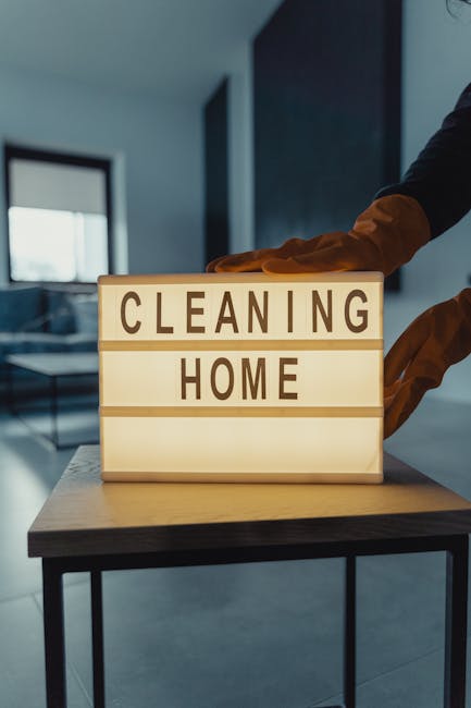 A person wearing orange rubber cleaning gloves is arranging or adjusting a lightbox sign on a wooden table that reads 'CLEANING HOME'. The background features a modern, well-lit living room with a gla